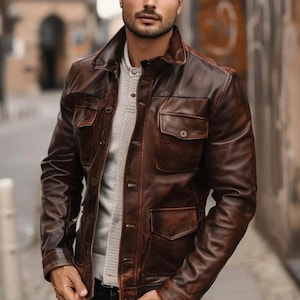 Puede incluir: Chaqueta de cuero marrón con un diseño clásico. La chaqueta presenta un cuello, cierre de botones y cuatro bolsillos delanteros. El cuero tiene un aspecto envejecido, con variaciones de color y textura.