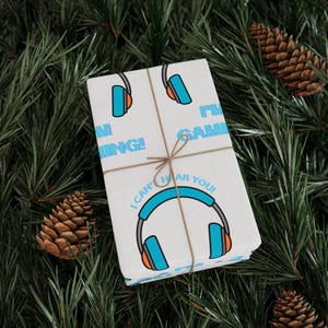 May include: A wrapped gift with white paper featuring a pattern of blue and orange headphones and the phrases "I'm Gaming!" and "I Can't Hear You!". The gift is tied with twine and set on a bed of pine needles and pine cones.