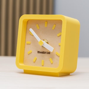 May include: A square yellow alarm clock with a light wood-tone face. The clock has a white minute and hour hand, and the text "WoodArt Lab" is printed on the face. The clock is on a light-colored surface.