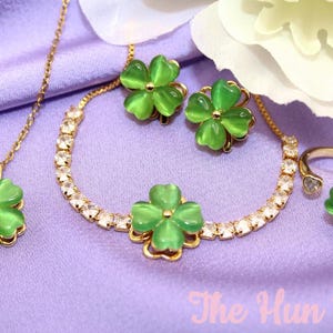 Op de afbeelding: Een goudkleurige sieradenset met klaverblad-bedels met groene kattenoogstenen. De set bevat een ketting, een tennisarmband, oorknopjes en een open ring. De tekst "The Hun Buns" is zichtbaar in de rechterbenedenhoek van de afbeelding.