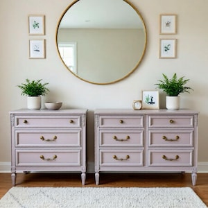 Puede incluir: Una escena de dormitorio con dos cómodas de color lila con herrajes de latón, un gran espejo redondo dorado y estampados botánicos enmarcados. La habitación está decorada con una alfombra blanca y helechos en macetas, creando un ambiente sereno y elegante.