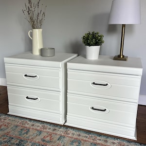 Puede incluir: Dos mesitas de noche blancas con dos cajones cada una y tiradores negros. Una tiene una lámpara con pantalla blanca y base dorada, la otra un jarrón con flores secas y un cuenco gris. Una alfombra está en primer plano. Las mesas son de madera.