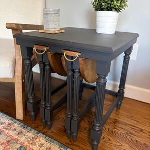 May include: A set of three nesting tables painted dark gray. The tables have a rectangular top, turned legs, and a wooden pull-out section with brass handles. A small potted plant and a glass with a coaster are on top.