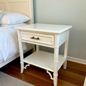 May include: A white bedside table with a drawer and a lower shelf. The table has a bamboo-style design with a dark bronze drawer pull. The table is next to a bed with white bedding.