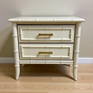 May include: Cream-coloured two-drawer bedside table with bamboo-style detailing. The drawers have gold-coloured handles and are outlined with a dark brown trim. The legs and frame mimic bamboo stalks, creating a cohesive design.