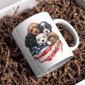 Op de afbeelding: Witte keramische mok met een ontwerp van vijf schattige puppy's in verschillende kleuren, die tegen elkaar aan liggen en gedrapeerd zijn met een Amerikaanse vlag. De mok heeft een wit handvat en staat tegen een achtergrond van bruin verpakkingsmateriaal.