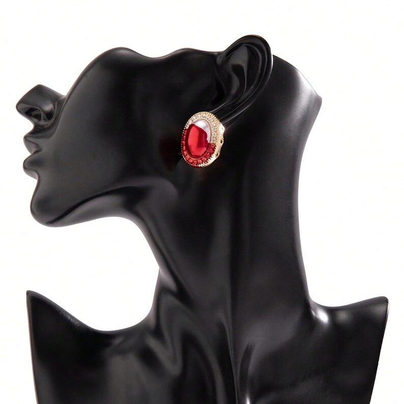 May include: A pair of gold-tone earrings with red oval-shaped stones set in a clear crystal halo.
