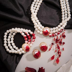 May include: A pearl necklace, bracelet, and earrings set with red and white accents. The necklace has a dangling charm with red beads.