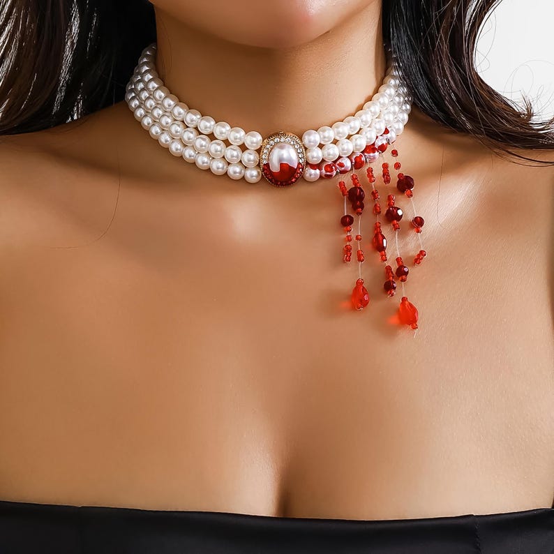 May include: A white pearl choker necklace with a red gemstone pendant and a dangling red beaded fringe.
