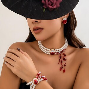 May include: A woman wearing a pearl necklace with a red gemstone pendant and a matching pearl bracelet with a red gemstone pendant. The necklace has a dangling strand of red beads.