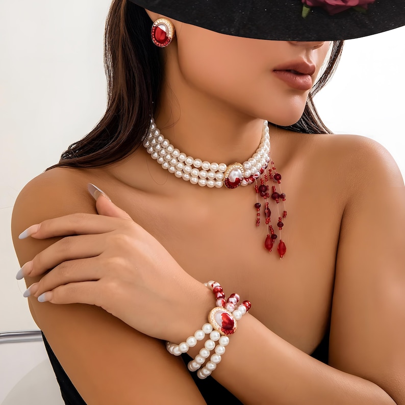 May include: A three-strand pearl choker necklace with a red and gold pendant and matching bracelet. The pendant features a large red gemstone surrounded by smaller red gemstones. The necklace and bracelet have a dangling red and gold charm.