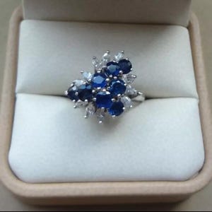 K&ouml;nnte beinhalten: Ein silberner Ring mit einem Cluster aus runden, tiefblauen Edelsteinen und kleineren, klaren, marquisef&ouml;rmigen Steinen. Der Ring wird in einer beigen Schmuckschatulle pr&auml;sentiert, die die funkelnde Anordnung der Edelsteine hervorhebt.