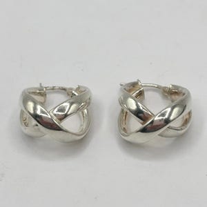 May include: A pair of silver-colored hoop earrings. Each earring features an intertwined design, creating an X shape. The earrings have a polished finish and a classic, minimalist style. The background is a plain, light color.
