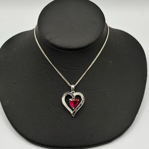 Puede incluir: Un collar de plata con un colgante en forma de corazón. El colgante presenta una gran piedra preciosa roja en el centro, rodeada por un borde de pequeñas piedras negras y blancas. El collar se exhibe en un maniquí negro.