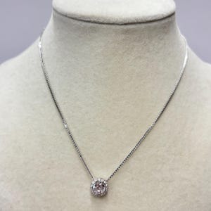 May include: A silver necklace featuring a clear, round gemstone pendant, encircled by smaller, clear stones. The necklace is presented on a mannequin.