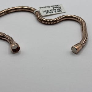 May include: A rose gold-plated bracelet with a flexible, ribbed design. The bracelet is made of .925 sterling silver and is plated with 14K rose gold. A small tag reads "925 S. Silver, 14K Rose Gold, Plated, Telesto Designs."