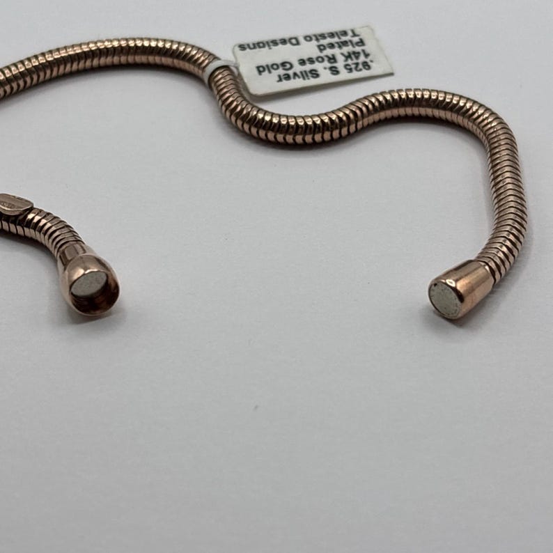 May include: A rose gold-plated bracelet with a flexible, ribbed design. The bracelet is made of .925 sterling silver and is plated with 14K rose gold. A small tag reads "925 S. Silver, 14K Rose Gold, Plated, Telesto Designs."