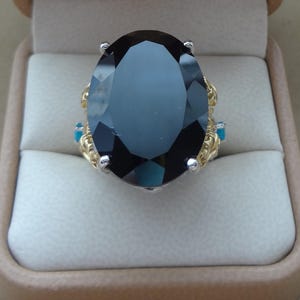 May include: A ring featuring a large, oval-cut black gemstone set in a silver-toned prong setting. The band is crafted from yellow gold-toned metal with intricate, textured detailing and small, bright blue accents on the sides of the setting.