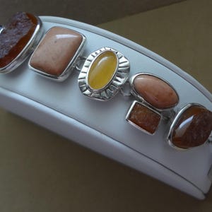 May include: A silver link bracelet featuring a series of multi-shaped gemstones in warm earth tones, including textured amber-colored stones, smooth peach-colored squares, and a central oval yellow stone set within a decorative silver frame.
