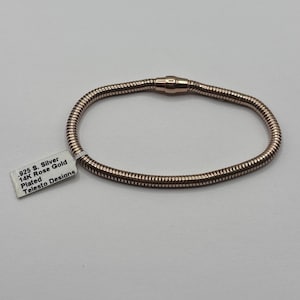 May include: A rose gold-plated bracelet with a magnetic clasp. The bracelet is made of a flexible, textured material. A white tag with the text ".925 S. Silver 14K Rose Gold Plated Telesto Designs" is attached.