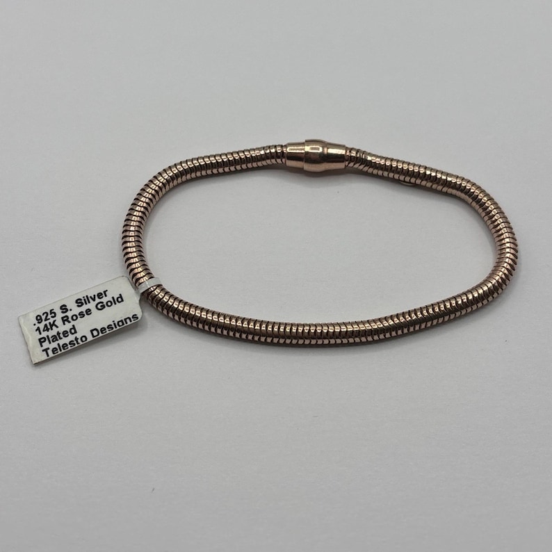 May include: A rose gold-plated bracelet with a magnetic clasp. The bracelet is made of a flexible, textured material. A white tag with the text ".925 S. Silver 14K Rose Gold Plated Telesto Designs" is attached.