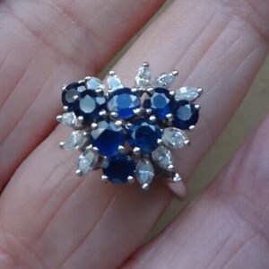 K&ouml;nnte beinhalten: Ein silberner Ring mit einem Cluster aus tiefblauen Saphiren, umgeben von kleineren, klaren Steinen in Marquiseform. Das Design des Rings &auml;hnelt einem Blumenarrangement, wobei die Saphire im Mittelpunkt stehen.