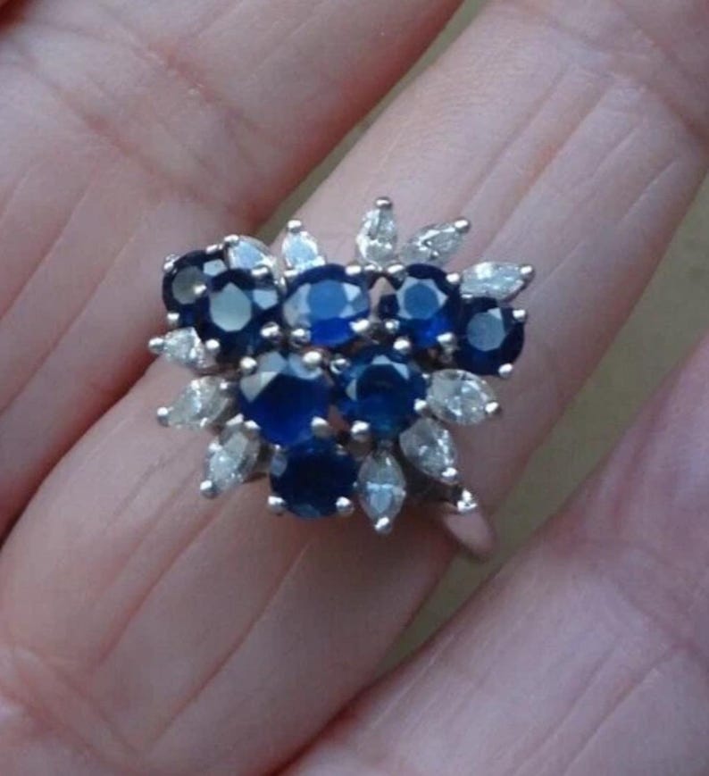K&ouml;nnte beinhalten: Ein silberner Ring mit einem Cluster aus tiefblauen Saphiren, umgeben von kleineren, klaren Steinen in Marquiseform. Das Design des Rings &auml;hnelt einem Blumenarrangement, wobei die Saphire im Mittelpunkt stehen.