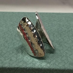 Puede incluir: Un anillo plateado con textura martillada. El anillo tiene un diseño único con dos secciones curvas superpuestas. La superficie refleja la luz, creando reflejos y sombras. El anillo se muestra sobre una superficie verde azulado.
