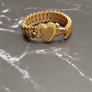 May include: A gold-toned expandable bracelet featuring a central heart-shaped charm with an engraved monogram. The band is constructed from a series of interlocking, textured metal links that create a flexible, continuous circular design.