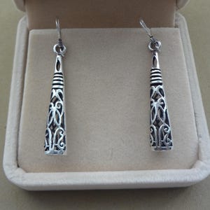 May include: A pair of silver-toned dangle earrings. Each earring has a conical shape with an open filigree design. The top portion features horizontal lines, and the earrings are displayed in a beige jewelry box.