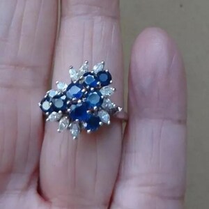 K&ouml;nnte beinhalten: Ein silberner Ring mit einem Cluster aus dunkelblauen und wei&szlig;en Edelsteinen. Das Design des Rings &auml;hnelt einem Blumenarrangement, wobei die blauen Steine den Mittelpunkt bilden und die wei&szlig;en Steine sie umgeben.