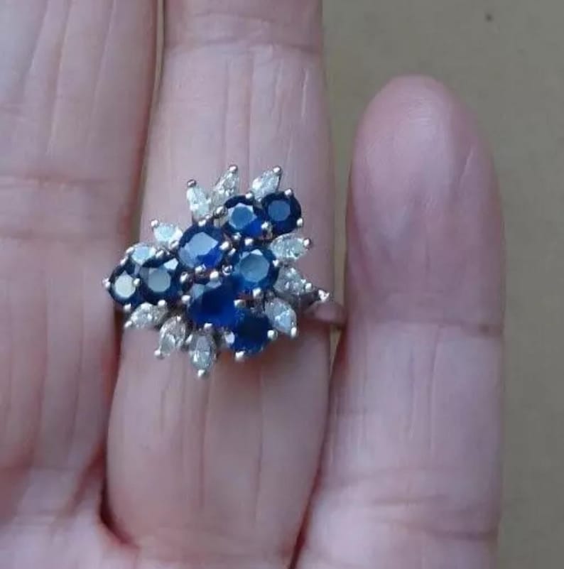 K&ouml;nnte beinhalten: Ein silberner Ring mit einem Cluster aus dunkelblauen und wei&szlig;en Edelsteinen. Das Design des Rings &auml;hnelt einem Blumenarrangement, wobei die blauen Steine den Mittelpunkt bilden und die wei&szlig;en Steine sie umgeben.