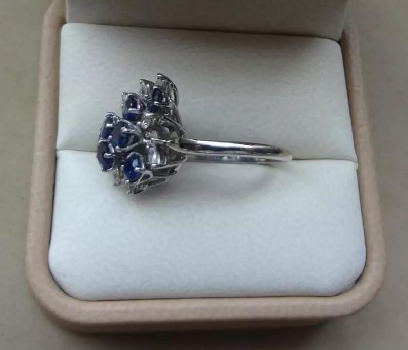 K&ouml;nnte beinhalten: Ein silberner Ring mit einem Cluster aus blauen Edelsteinen. Der Ring hat ein Design mit mehreren blauen Steinen in einem Blumen- oder Sternmuster. Der Ring wird in einer beigen Schmuckschatulle pr&auml;sentiert.