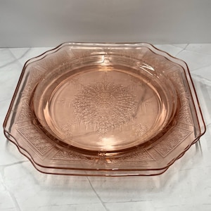 May include: A vintage pink glass plate with an octagonal shape and a detailed floral and geometric pattern. The plate has a raised rim and a central design, perfect for serving or display.