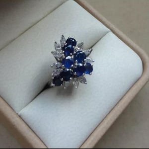 K&ouml;nnte beinhalten: Ein silberner Ring mit einem Cluster aus dunkelblauen und wei&szlig;en Edelsteinen. Die Edelsteine sind in einem floralen Muster angeordnet, mit den dunkelblauen Steinen in der Mitte und wei&szlig;en Steinen, die sie umgeben. Der Ring befindet sich in einer hellen Schachtel.
