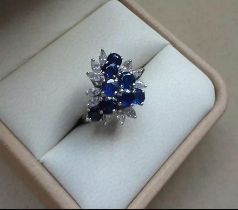 K&ouml;nnte beinhalten: Ein silberner Ring mit einem Cluster aus dunkelblauen und wei&szlig;en Edelsteinen. Die Edelsteine sind in einem floralen Muster angeordnet, mit den dunkelblauen Steinen in der Mitte und wei&szlig;en Steinen, die sie umgeben. Der Ring befindet sich in einer hellen Schachtel.