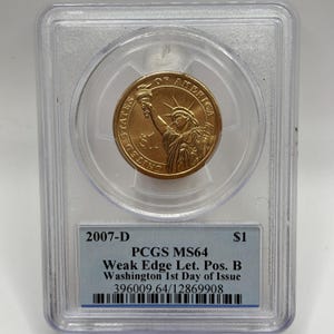 May include: A gold-coloured $1 coin featuring the Statue of Liberty, encased in a clear plastic holder. The coin is labelled "2007-D" and "PCGS MS64." The holder also includes text indicating it is a "Washington 1st Day of Issue" coin.