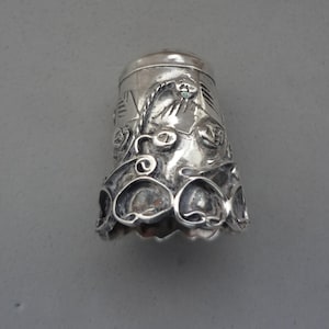 Puede incluir: Un dedal de plata con intrincados diseños florales y de volutas. El dedal tiene una base acampanada con recortes en forma de corazón y una parte superior detallada. El metal tiene un acabado pulido. Un accesorio de costura.