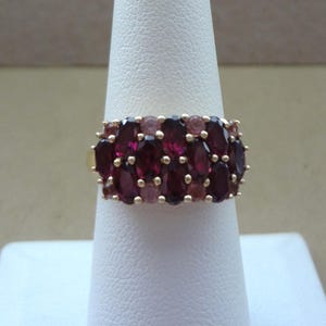 Könnte beinhalten: Ein goldener Ring mit einem Cluster aus oval geschliffenen, tiefroten Edelsteinen, akzentuiert mit kleineren rosa Edelsteinen. Der Ring wird auf einem weißen Ringhalter präsentiert, der das Design und den Farbkontrast hervorhebt.