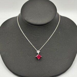 Puede incluir: Un collar de plata con un pequeño colgante cuadrado de piedra preciosa rojo rubí. Encima de la piedra roja hay una piedra más pequeña, blanca, en forma de diamante. El collar se exhibe en un busto de joyería negro.