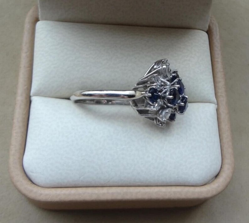 K&ouml;nnte beinhalten: Ein silberner Ring mit einem Cluster aus dunkelblauen und klaren Edelsteinen. Der Ring hat ein filigranes Design und wird in einer beigen Ringschatulle pr&auml;sentiert.