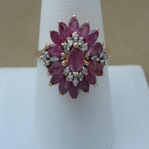 Könnte beinhalten: Ein goldener Ring mit einem floralen Design, besetzt mit tiefroten, ovalen Edelsteinen und kleinen, klaren Edelsteinen. Der Ring wird auf einer weißen Oberfläche präsentiert. Die roten Edelsteine sind blütenförmig angeordnet.