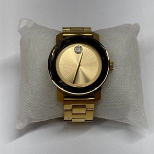May include: A gold-toned Movado watch with a black bezel and a single diamond at the 12 o'clock position. The watch has a gold-toned metal strap.