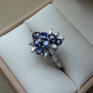 K&ouml;nnte beinhalten: Ein silberner Ring mit einem bl&uuml;tenf&ouml;rmigen Design, besetzt mit dunkelblauen Edelsteinen und kleineren wei&szlig;en Steinen. Der Ring liegt in einer beigen Schatulle, die den Kontrast zwischen dem Schmuck und dem Hintergrund hervorhebt.