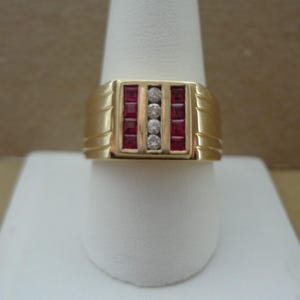 May include: A gold ring with a rectangular face. The face features three vertical sections: two with red stones and a center section with three small, round, clear stones. The ring is displayed on a white stand.