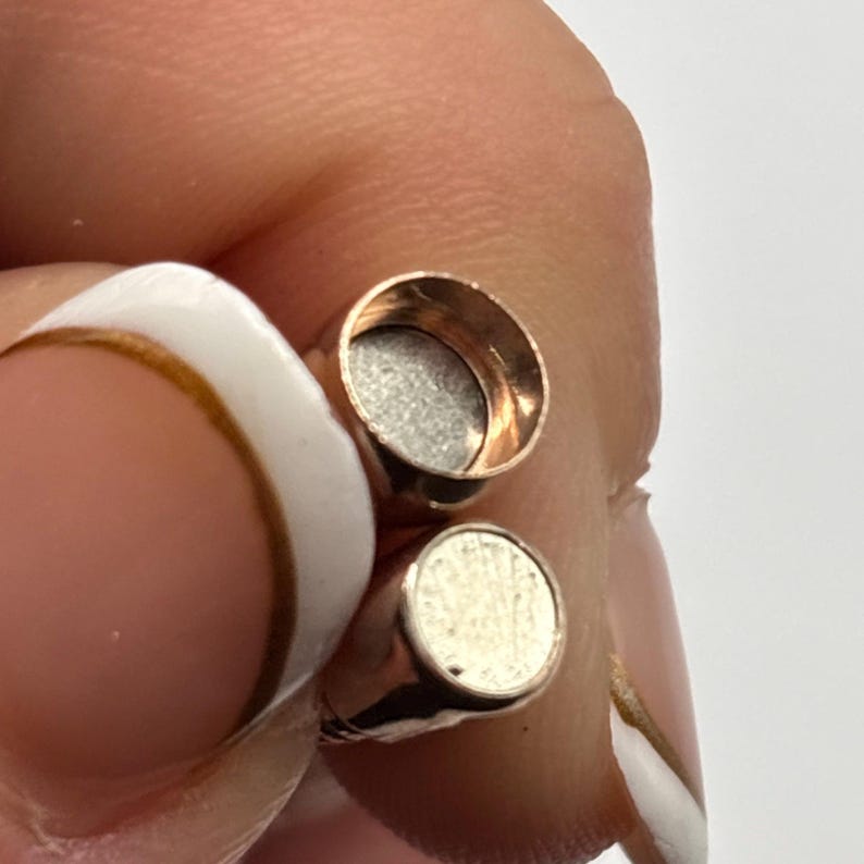 May include: Two small, rose gold-colored magnetic clasps. Each clasp has a circular, flat, silver-colored magnet on one side. The clasps are designed to connect magnetically. The clasps are shown against a light background.