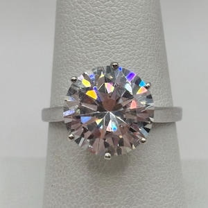Op de afbeelding: Een ring met een grote, ronde, heldere edelsteen en een zilveren band. De edelsteen is facetgeslepen en vangt het licht op, waarbij een regenboog aan kleuren wordt gereflecteerd. De ring staat op een witte achtergrond.