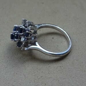 K&ouml;nnte beinhalten: Ein silberner Ring mit einem Cluster aus dunkelblauen Edelsteinen. Die Edelsteine sind in einem mehrschichtigen Design angeordnet, das einem Blumen- oder Sternausbruch &auml;hnelt. Der Ring hat einen glatten, polierten Ring.