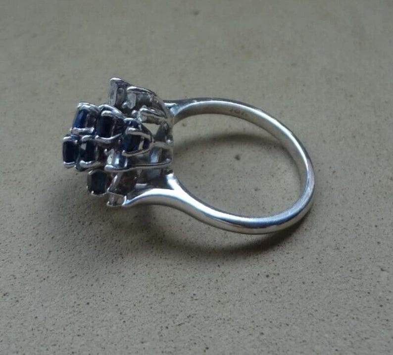 K&ouml;nnte beinhalten: Ein silberner Ring mit einem Cluster aus dunkelblauen Edelsteinen. Die Edelsteine sind in einem mehrschichtigen Design angeordnet, das einem Blumen- oder Sternausbruch &auml;hnelt. Der Ring hat einen glatten, polierten Ring.