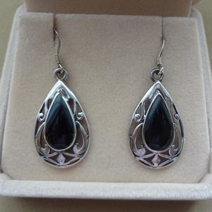 May include: A pair of silver drop earrings featuring a central smooth black teardrop-shaped stone encased in an intricate openwork metal frame with scroll details, suspended from simple silver ear wires for a secure and balanced appearance.
